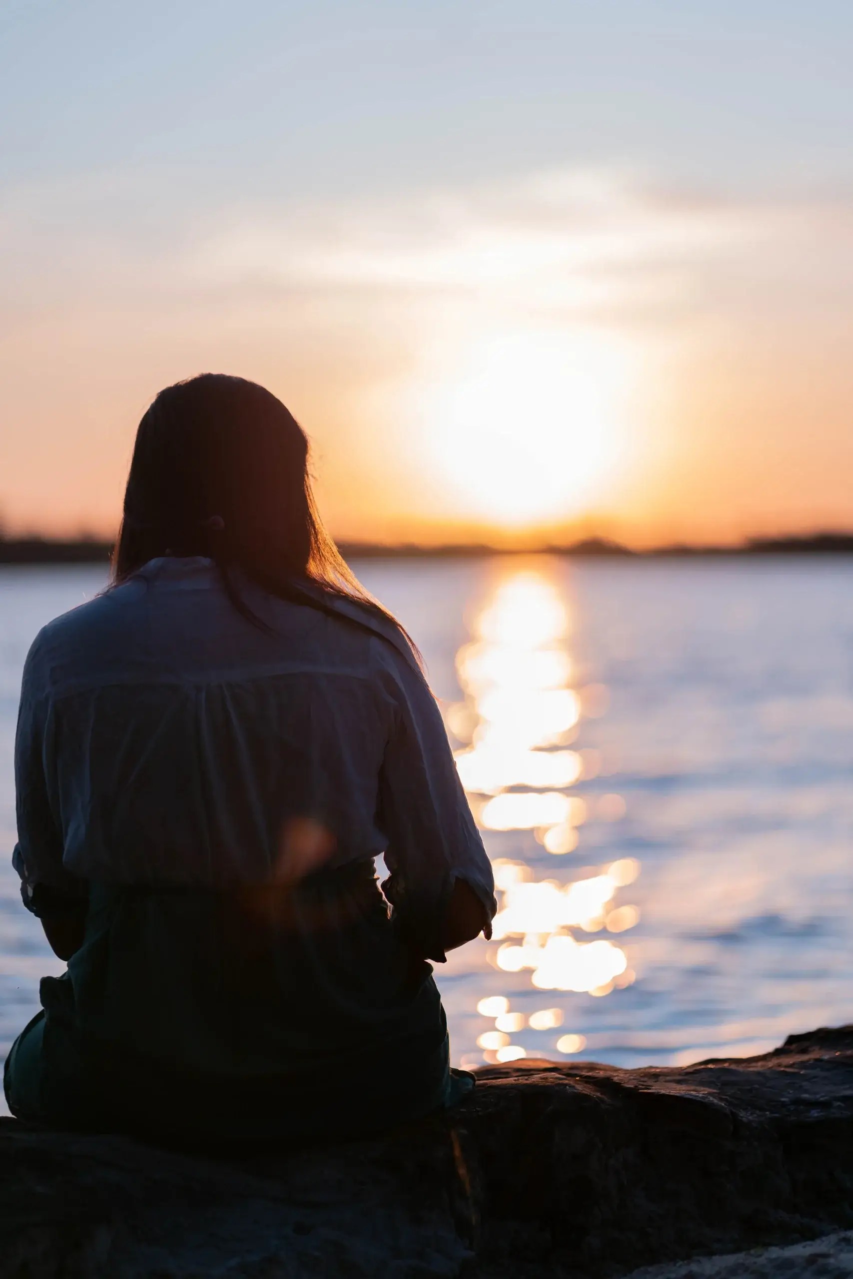 A woman sitting beside the water and watching the sunset.