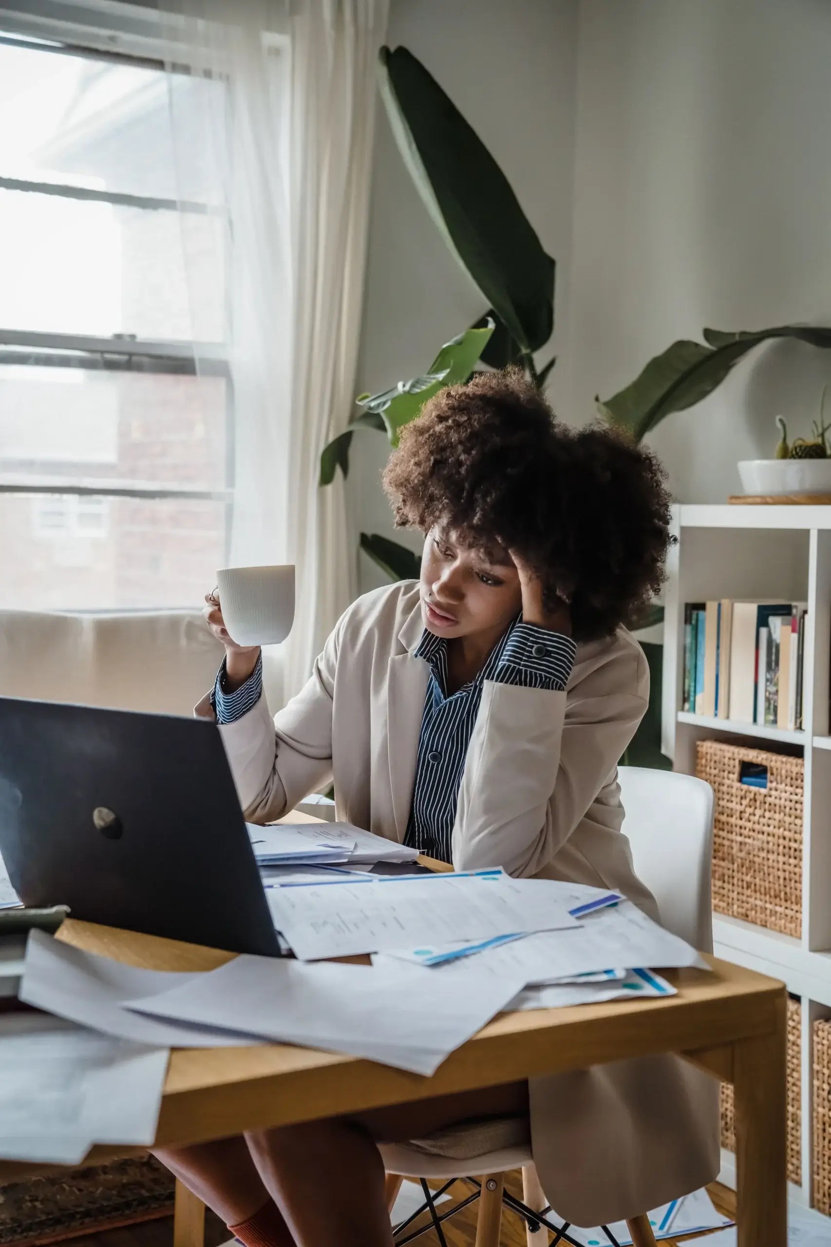 Stressed woman sitting at a desk with paperwork, holding a cup and resting her head on her hand