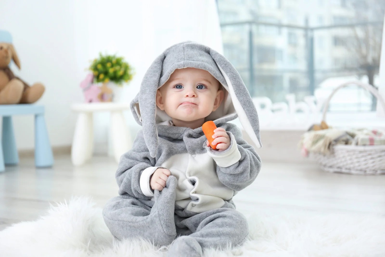 Cute little baby in bunny costume sitting on furry rug and eating carrot