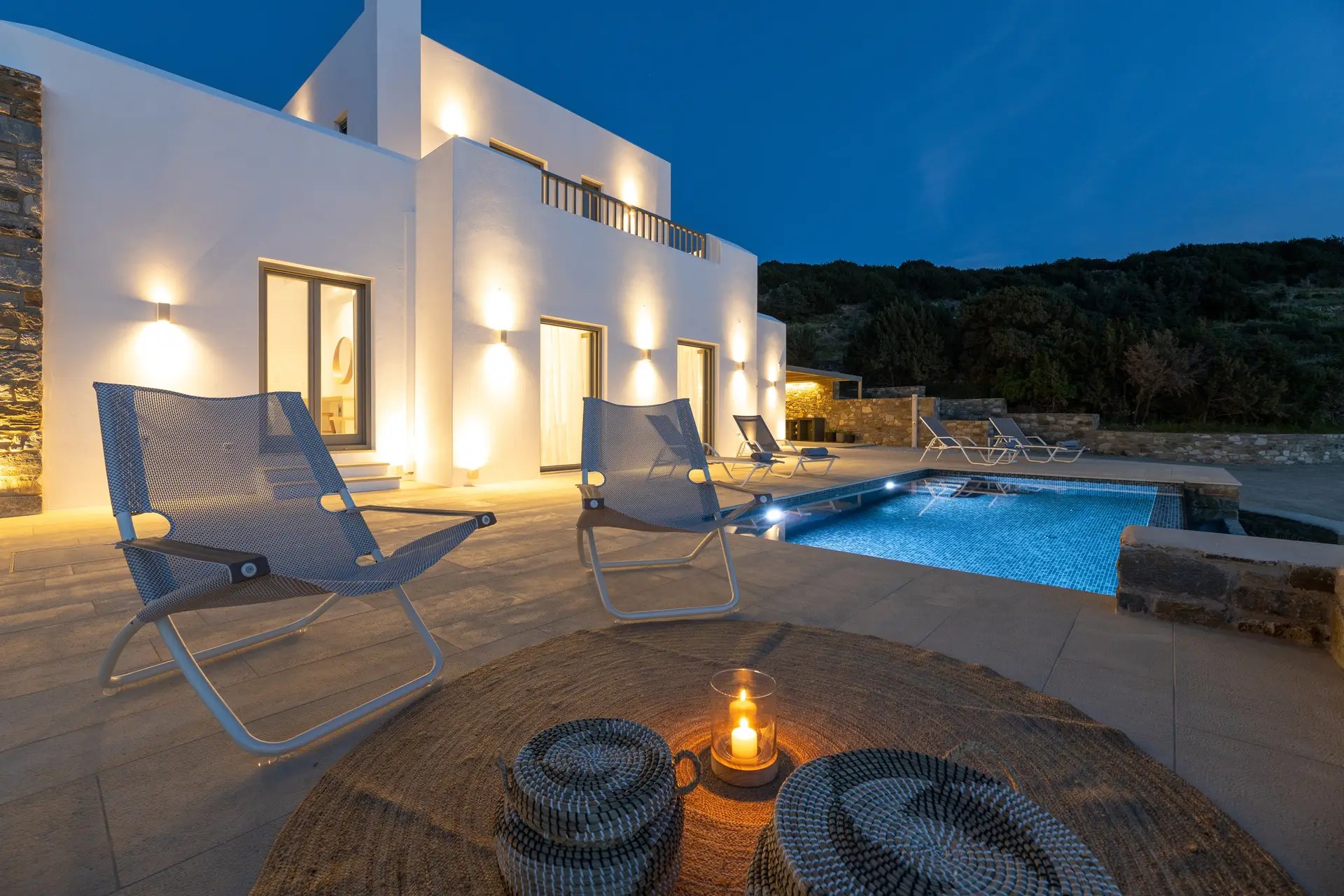 Evening poolside at a luxury Paros villa with illuminated outdoor lounge, cozy loungers, and candlelit decor for a serene ambiance.