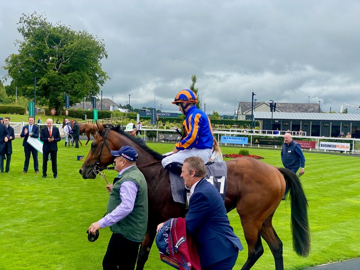 A jockey in bright orange and blue attire riding a horse, being led by a man on the ground. Several spectators and officials are in the background, with a grassy racetrack and cloudy skies.