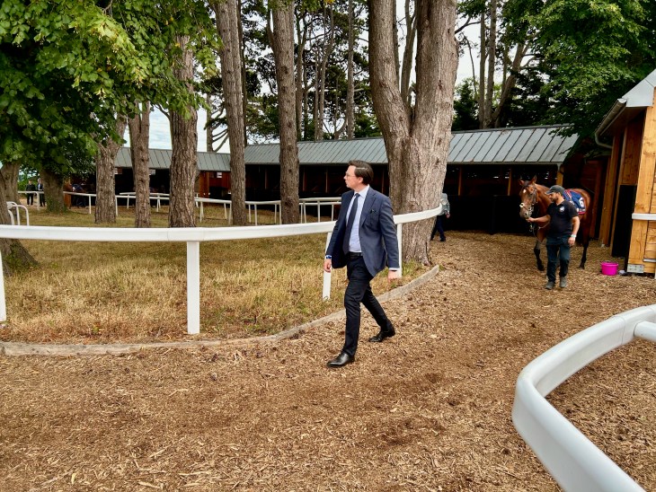 A man in a suit walking along a path in a horse stable area with trees and stables in the background.