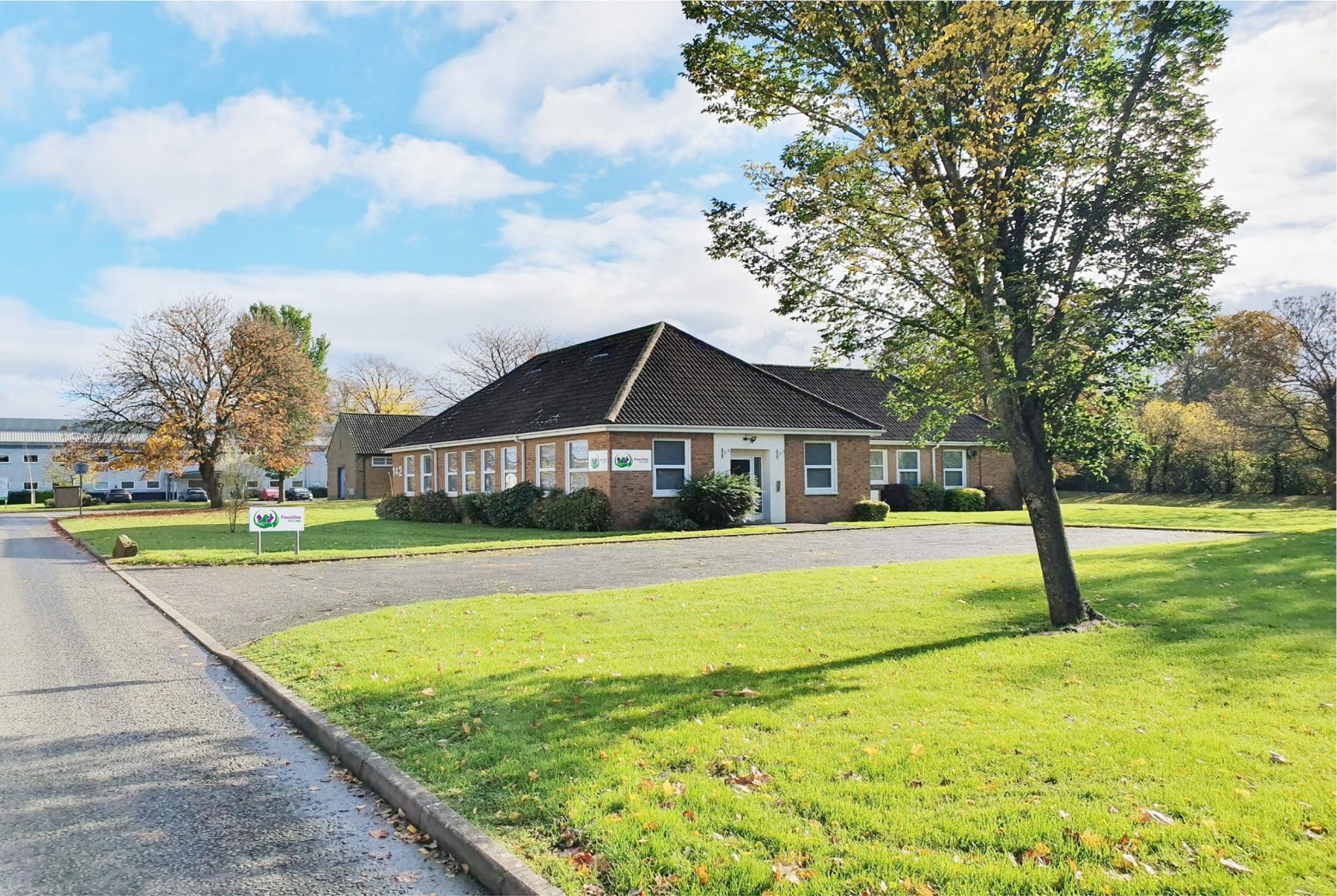 Pawsitive Vet Care's premises, a single storey brick building with sloping roof with bushes around the side, a tarmacadam car park in front and grass with a tree in the foreground