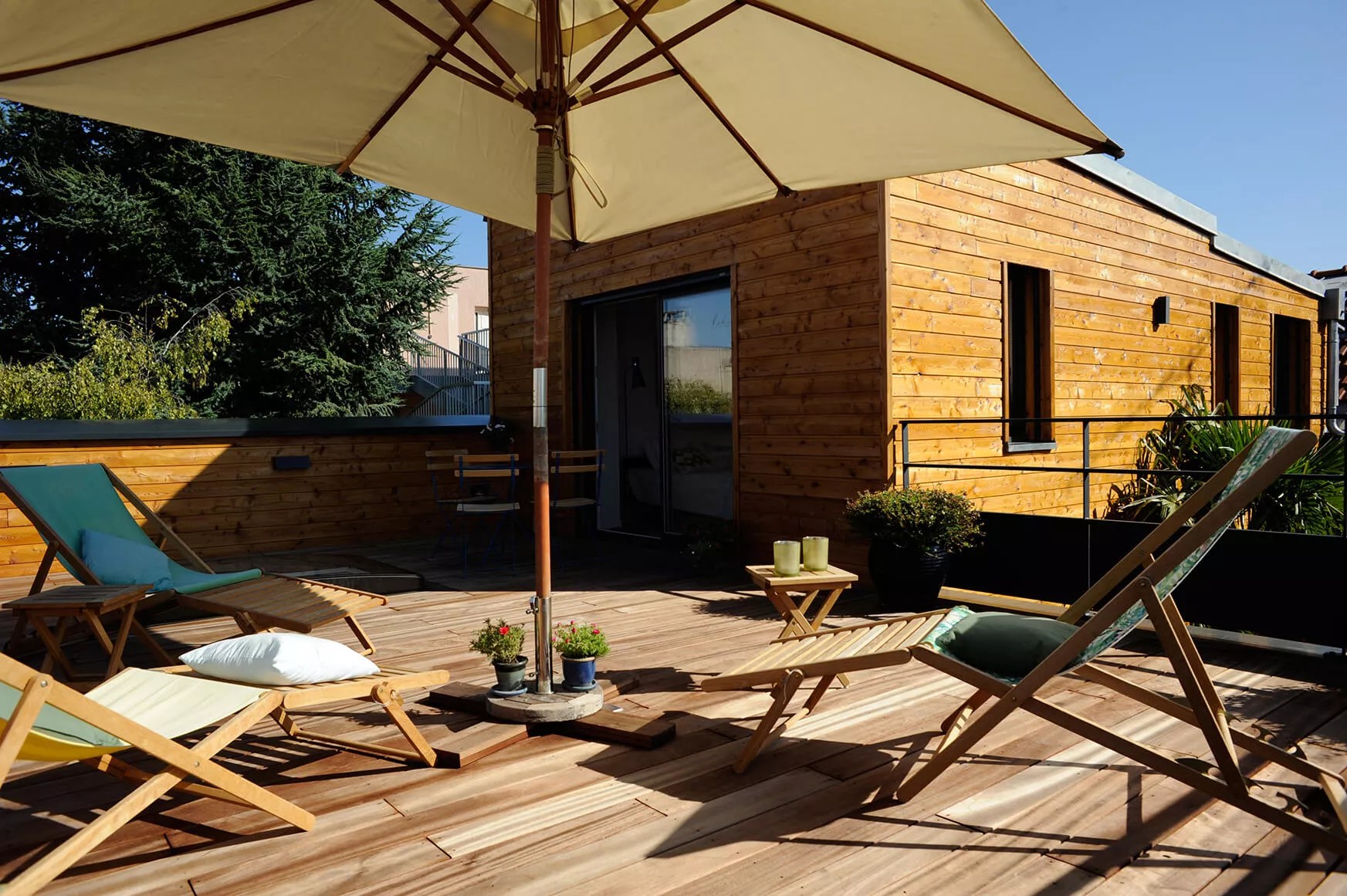 Maison vue de l'exterieur en été, avec terrasse en bois, parasol et végétation