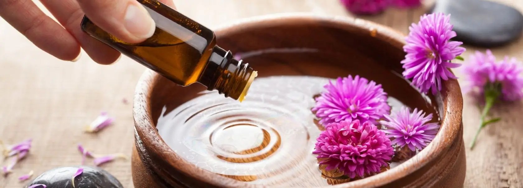 Hand pouring essential oil from amber glass bottle into a wooden bowl of water with floating purple flowers, surrounded by smooth black stones and scattered petals—aromatherapy ritual for relaxation and holistic wellness.