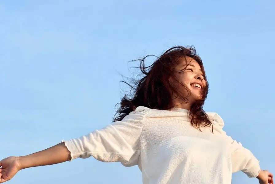 Woman with long dark hair smiling with arms outstretched under a clear blue sky, wearing a white top. Outdoor scene evokes joy, freedom, optimism, and emotional clarity—ideal for mindfulness, mental wellness, and intentional living themes.