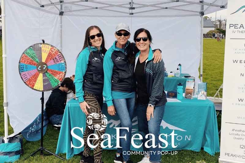 Three smiling female employees of Peak Hydration pose for a photo at the Peak Hydration tent at SeaFeast Bellingham