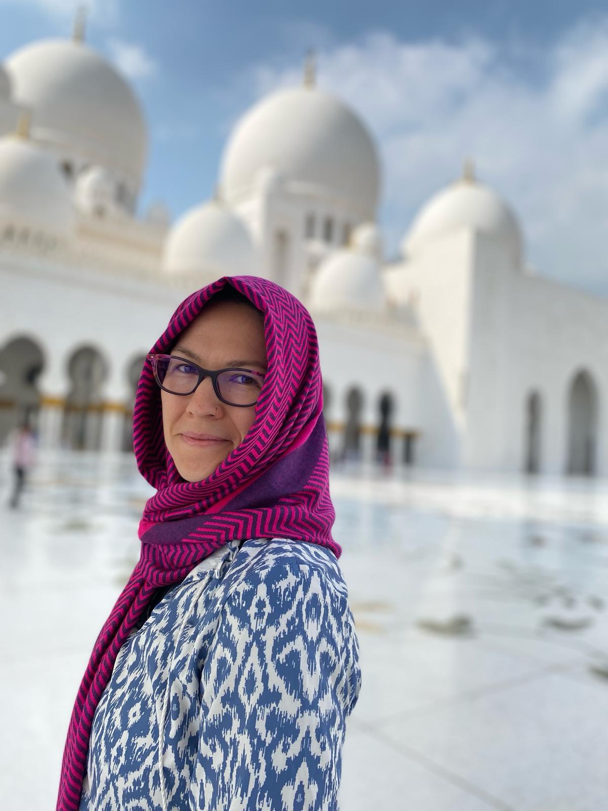 A women in a pink scarf and blue dress smiling with the white Sheikh Zayed Grand Mosque Center Abu Dhabi