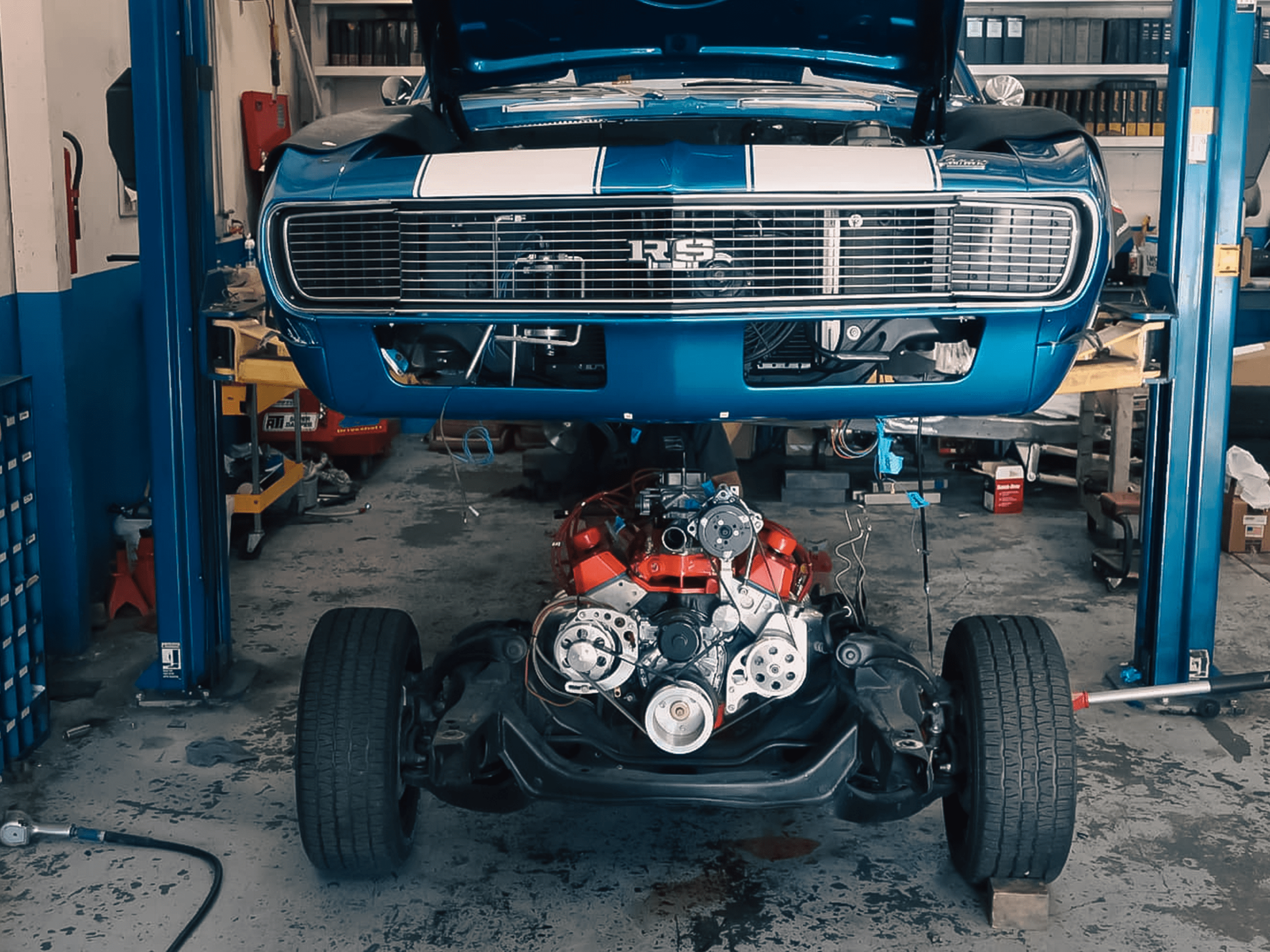 A blue Chevrolet Camaro RS raised on a hydraulic lift from the front, with a red high-performance V8 engine and drivetrain components visible beneath it.