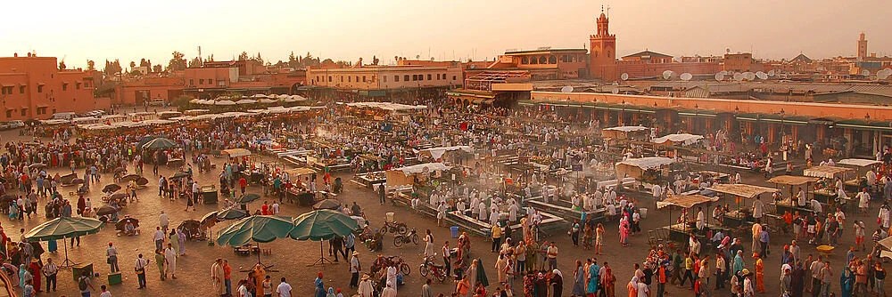 Busy open-air market square at sunset with vendors, tables, and crowds under umbrellas and awnings.