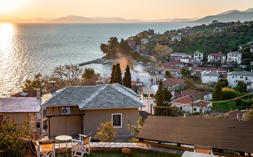 Sunset over a coastal village with hillside houses facing a calm bay to the left.