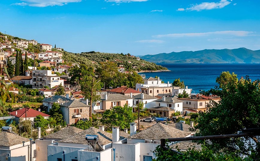 Coastal hillside town with white and pastel houses, narrow streets, and a blue sea beneath a clear sky.