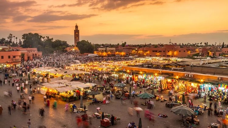 Bustling evening market square at sunset with crowded stalls and warm lights, Marrakech, with the Koutoubia Mosque minaret in the background.