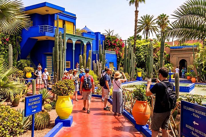 Tourists stroll along a red path beside cobalt-blue buildings, tall cacti, and colorful planters at Jardin Majorelle.