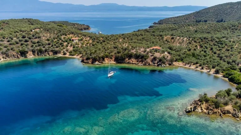 Aerial view of a turquoise bay with a solitary sailboat anchored near a rocky, tree-covered coastline in clear, calm water.