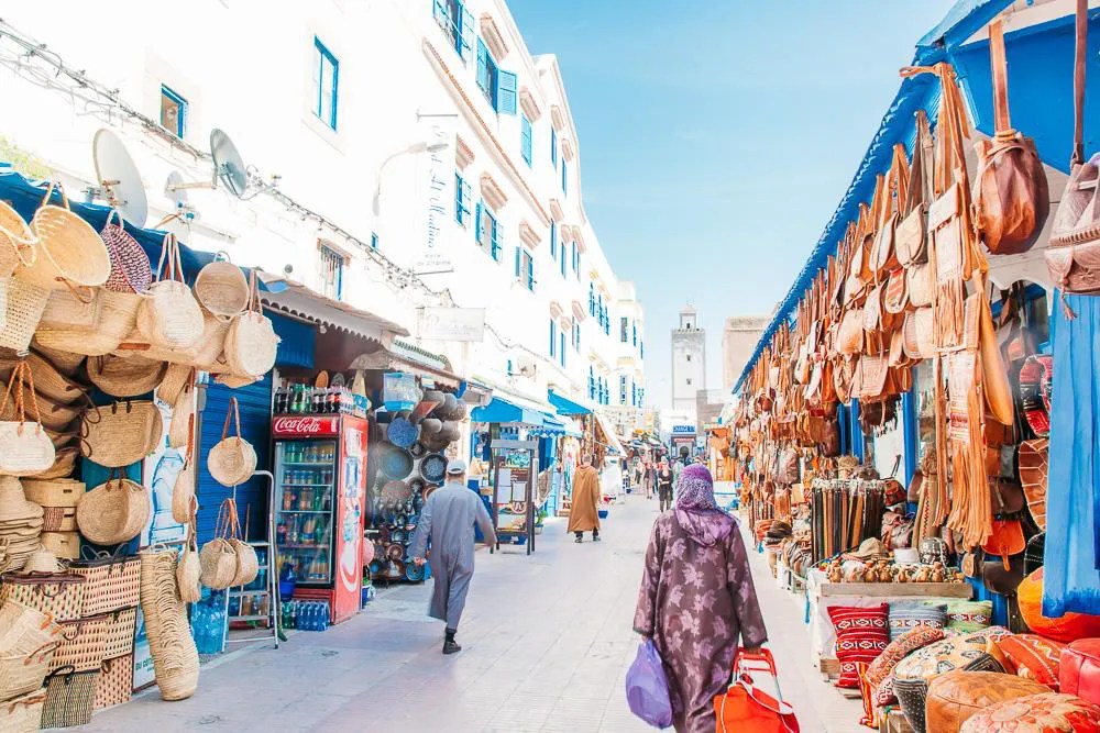 Bustling outdoor market street with woven baskets and leather goods for sale along white-and-blue buildings under a sunny sky.