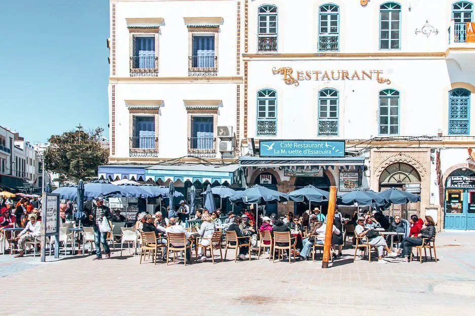 Outdoor cafe with many diners under blue umbrellas in a sunny plaza in front of a white building marked 'Restaurant' on a warm day