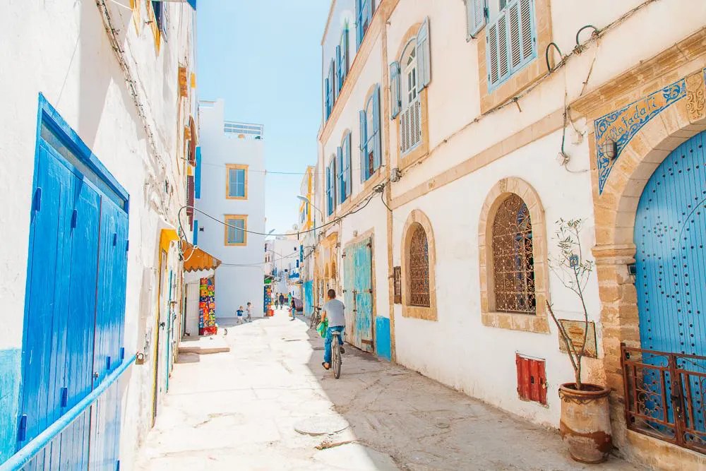 Narrow sunlit street in a white-washed town with blue doors and shutters; a person rides a bicycle down the lane.