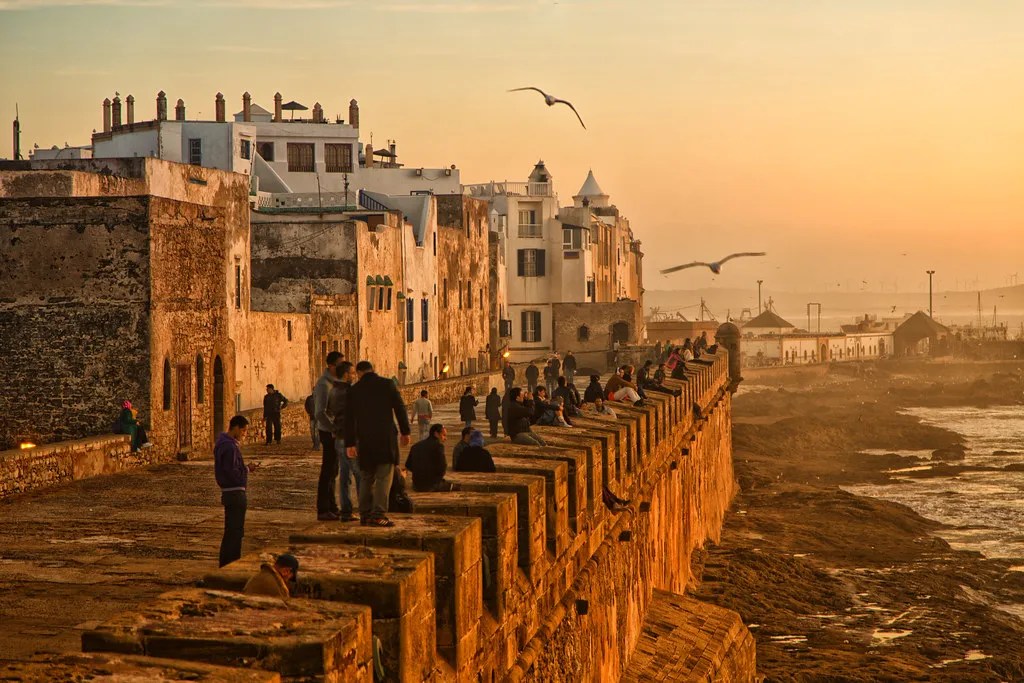 People walk along a stone seawall at sunset with white buildings and seagulls in the sky informed image is a coastal fortress sunset scene