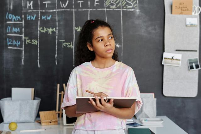 A teenager in a pink shirt ponders while holding a notebook in a classroom setting.