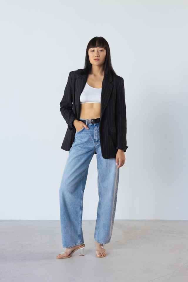 A stylish woman in a black blazer and jeans poses confidently in a studio setting.