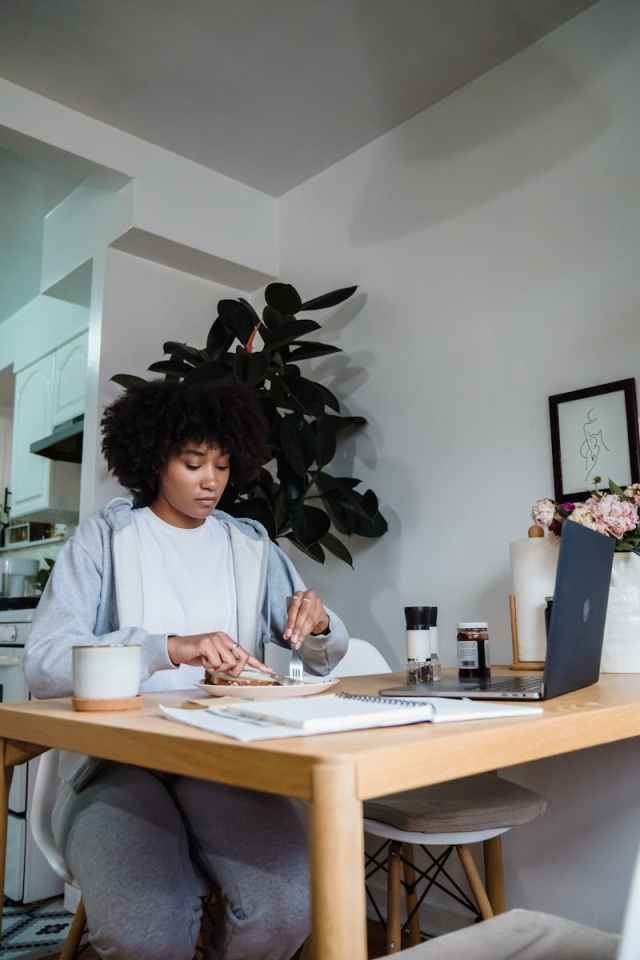 Young woman multitasking at home, working on laptop while enjoying a meal in the kitchen.
