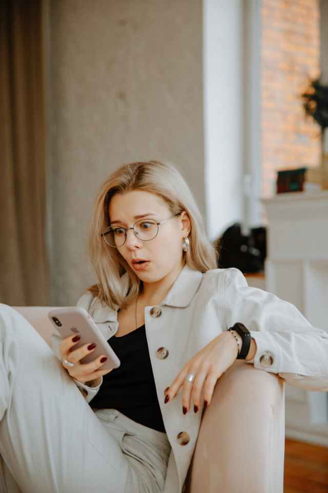 Surprised woman with glasses reacts to news on her phone while relaxing indoors.