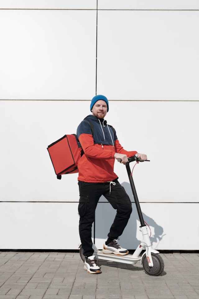 Young man on electric scooter with delivery bag, posing outdoors.