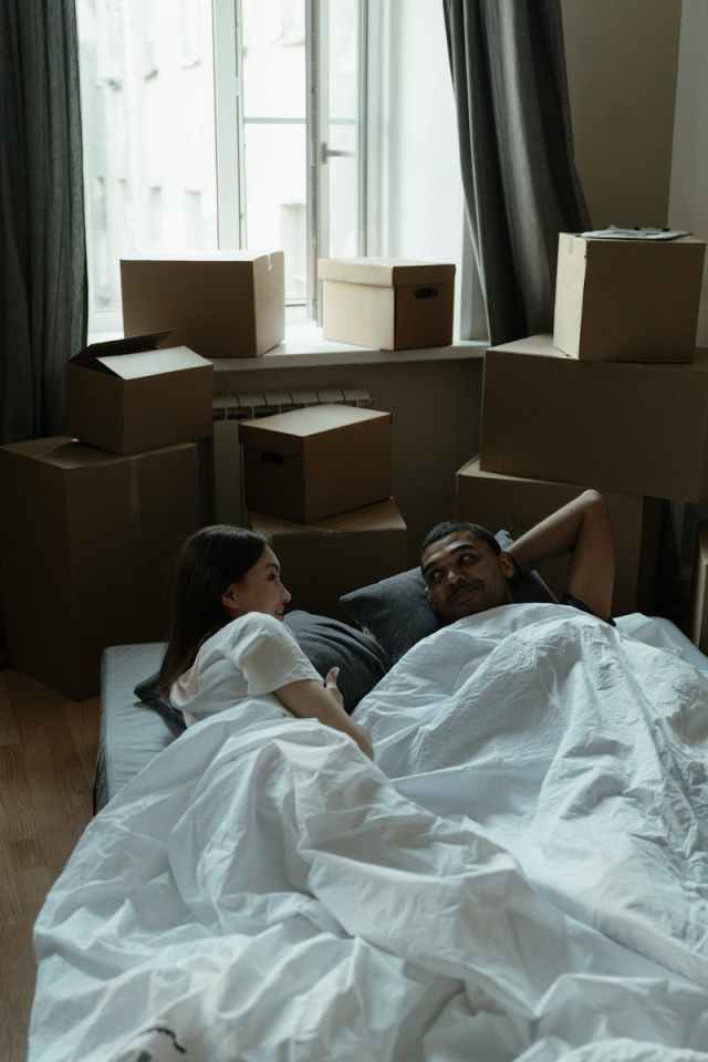A couple lies comfortably in bed surrounded by moving boxes in their new apartment.