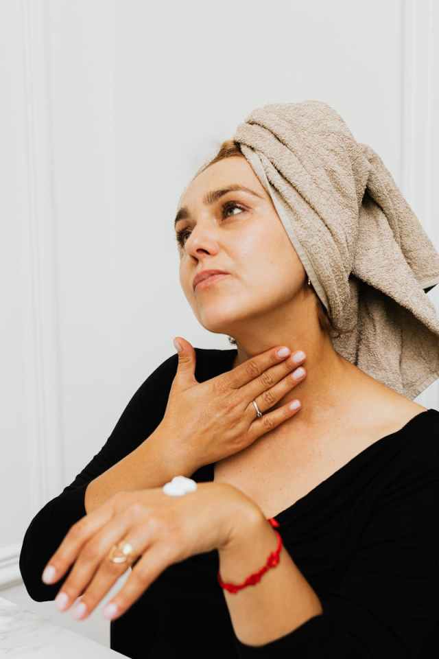 Woman with a towel on her head applying lotion to her neck post-bath.
