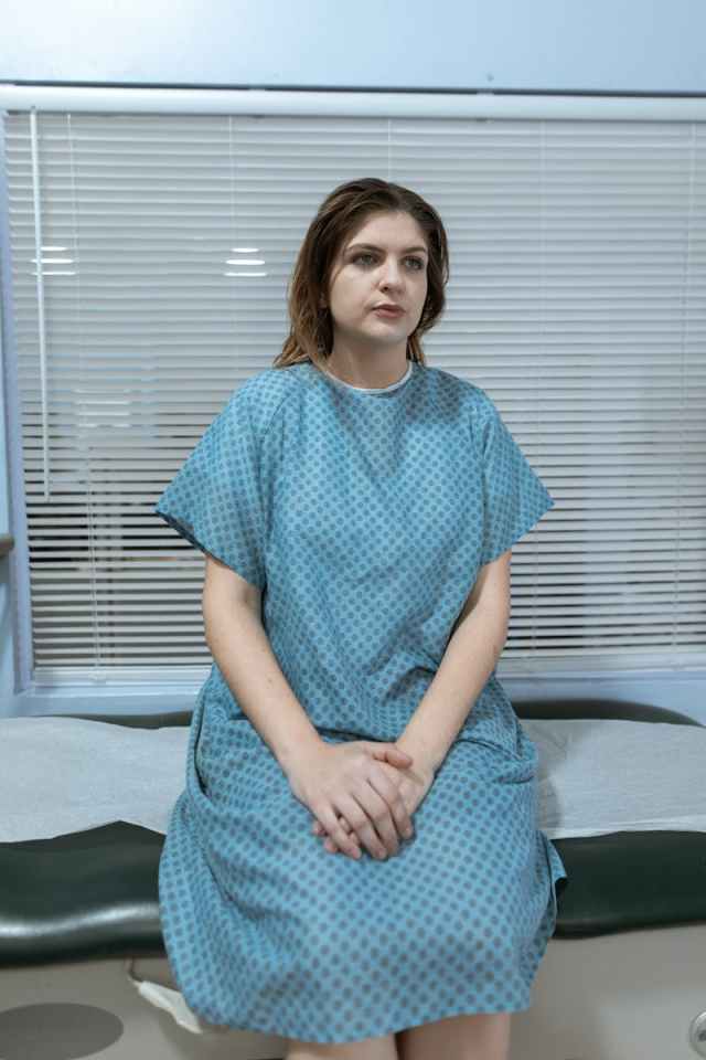 A woman in a hospital gown sitting on an exam table, looking contemplative.