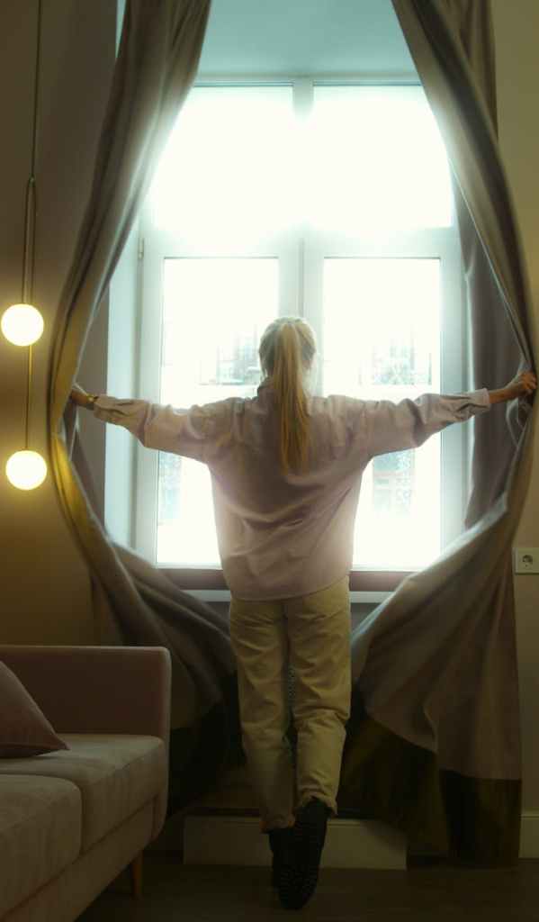 A serene woman opens curtains, welcoming natural light into a cozy room setting.