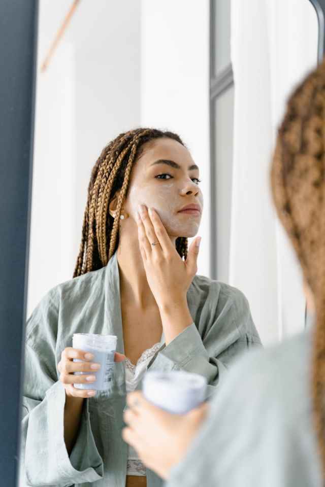 Woman in a green robe applying facial cream in front of the mirror for skin care.
