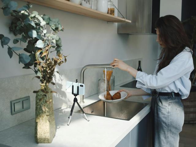 A woman washes pears under a faucet in a modern kitchen while recording with her phone.