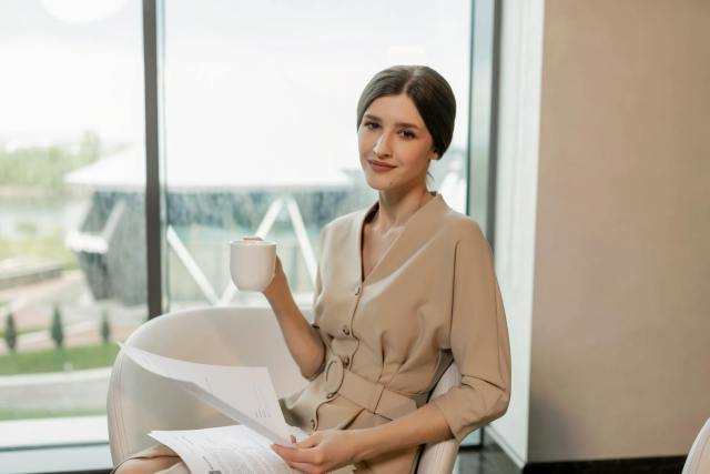A confident businesswoman enjoys coffee while sitting in a modern office setting.
