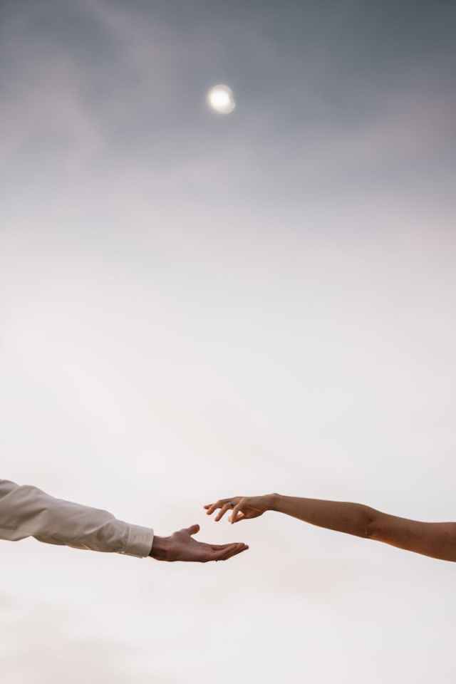 A man and a woman reaching out their hands towards each other under a serene sky.