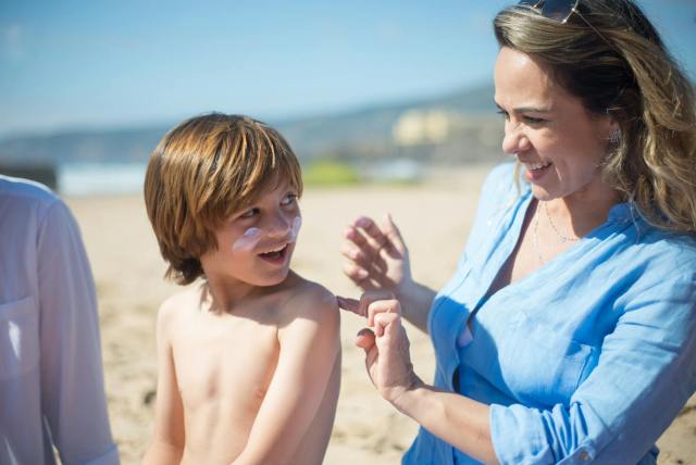 A woman applying sunscreen to her smiling son on a sunny beach day, promoting skin care and safety.
