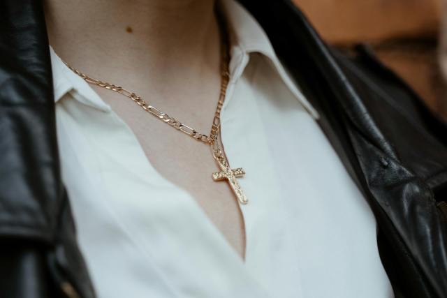 Close-up of a gold cross pendant necklace worn over a white blouse and leather jacket.