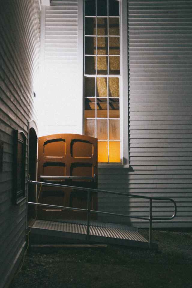 Atmospheric night image of a building entrance with a wooden door and metal ramp.