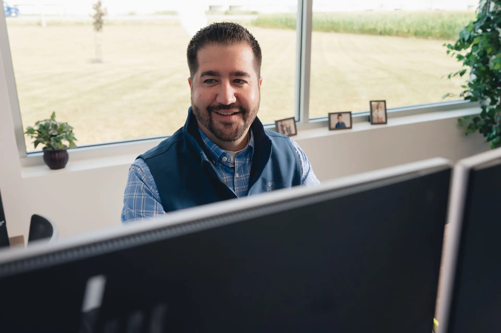 A bearded man smiles while working at a desk with dual monitors in a bright Ohio office. Framed photos and a potted plant sit on the windowsill behind him, with business technology tools set against a large window overlooking a grassy field.