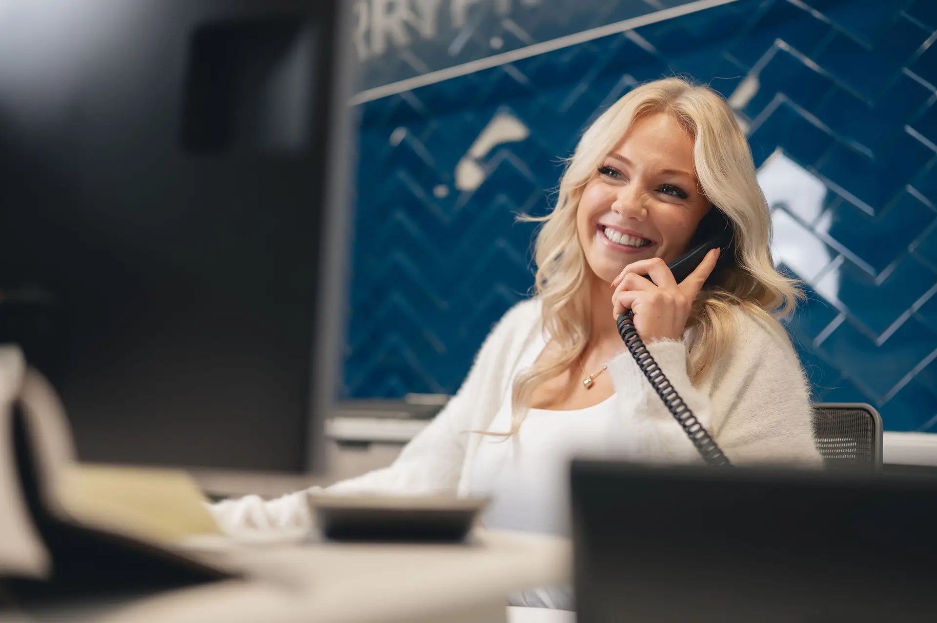 A woman with blonde hair smiles while talking on a corded phone at her desk in an office, discussing document management Ohio, with a blue tiled wall in the background.