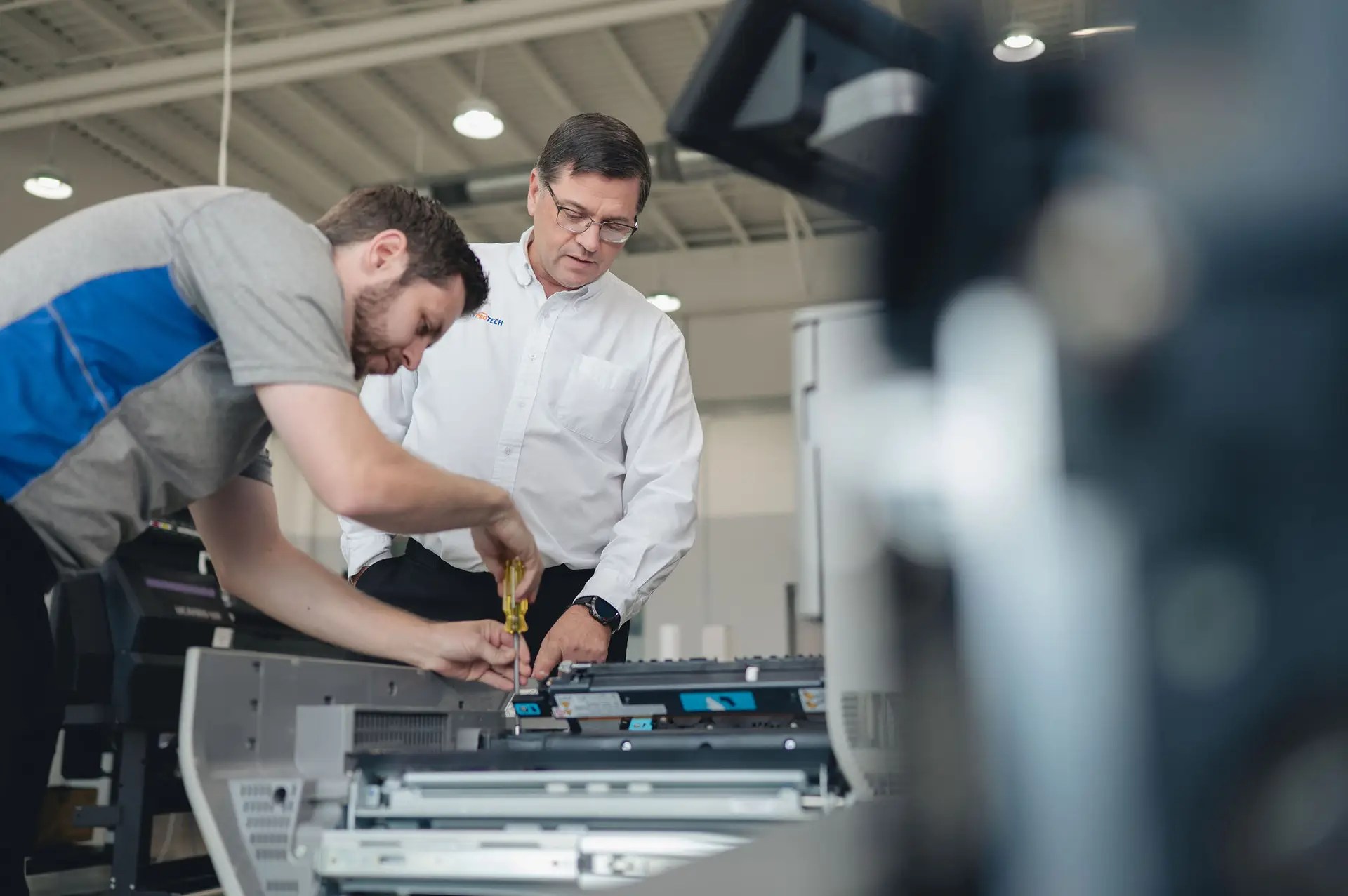 Two men working together in a modern industrial setting, one using a screwdriver to fix equipment while the other observes and gives guidance. Machinery and tools are visible in the foreground and background.