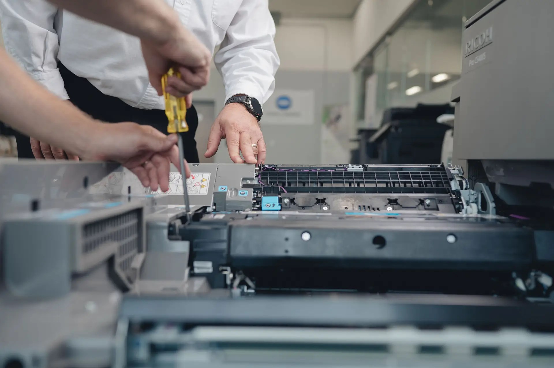 Two people repair a large office printer; one uses a screwdriver while the other supervises. The printers top is open, exposing internal components. The setting appears to be a modern office or workshop.