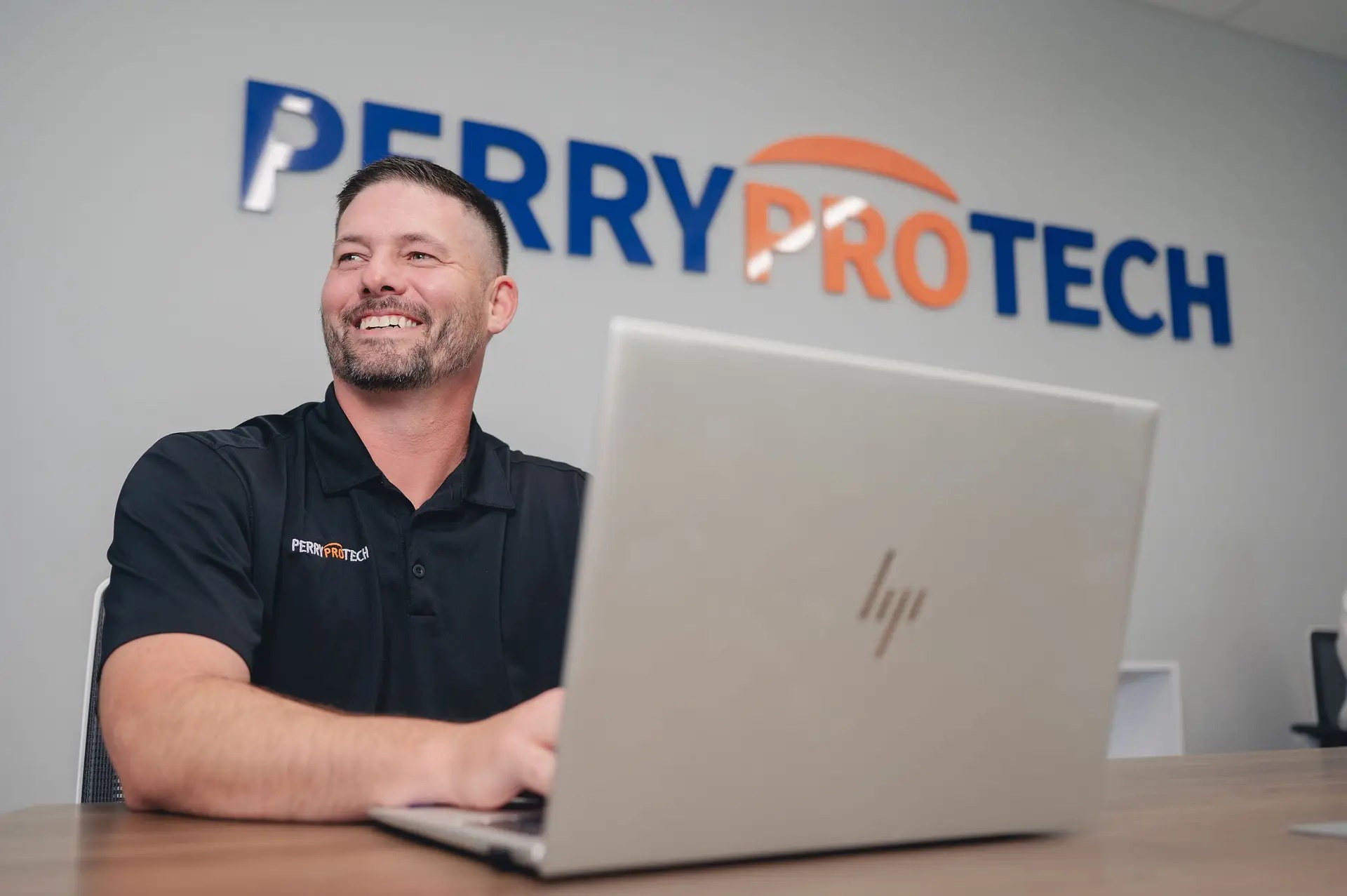 A man in a black Perry ProTech shirt smiles while working on a laptop at a desk, representing Managed IT and Network Services. The Perry ProTech company logo is visible on the wall behind him.