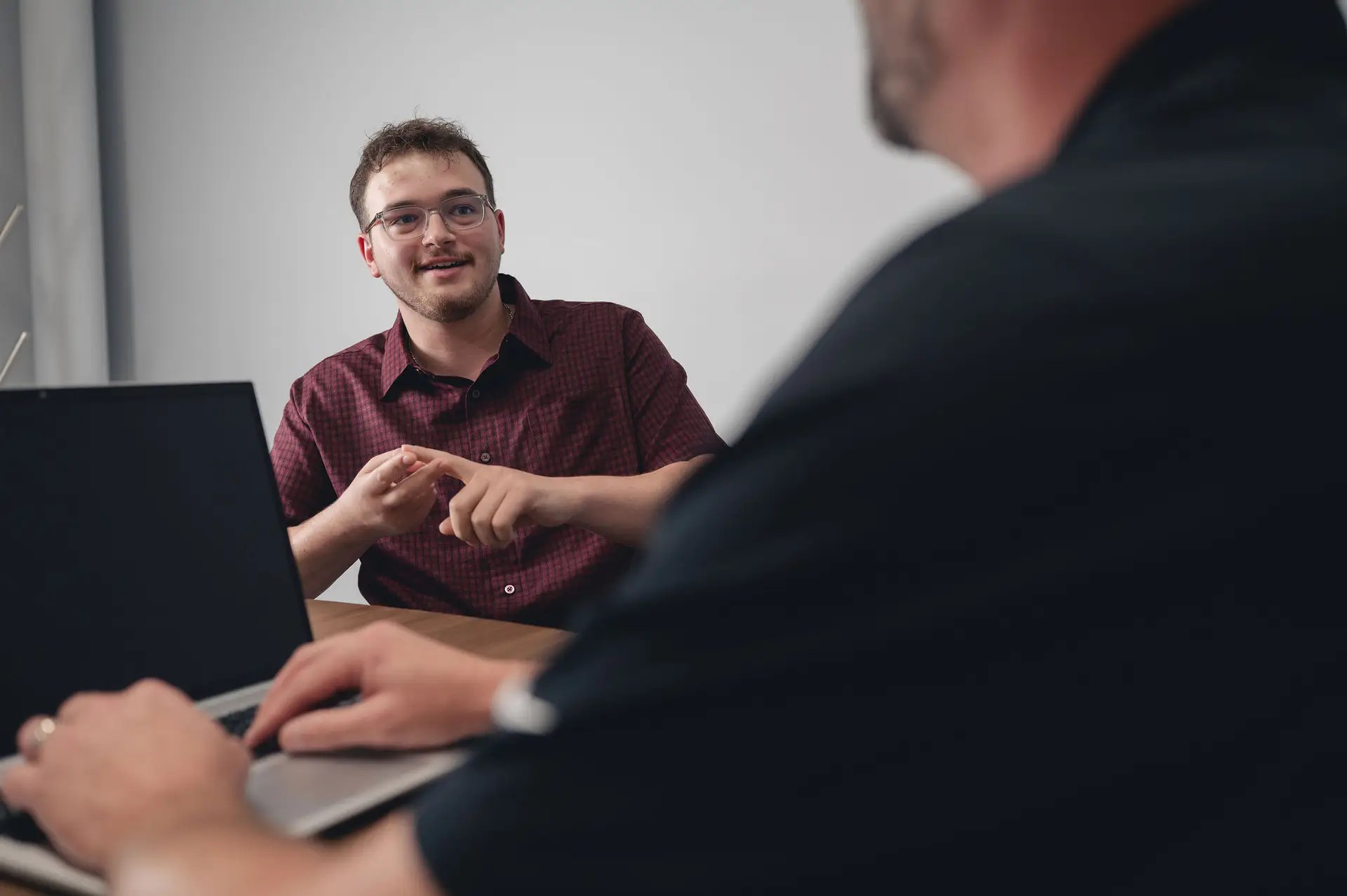 Two people are having a conversation at a desk. One person in the foreground is using a laptop, while the other gestures with their hands and appears to be speaking. The setting looks like an office or meeting room.