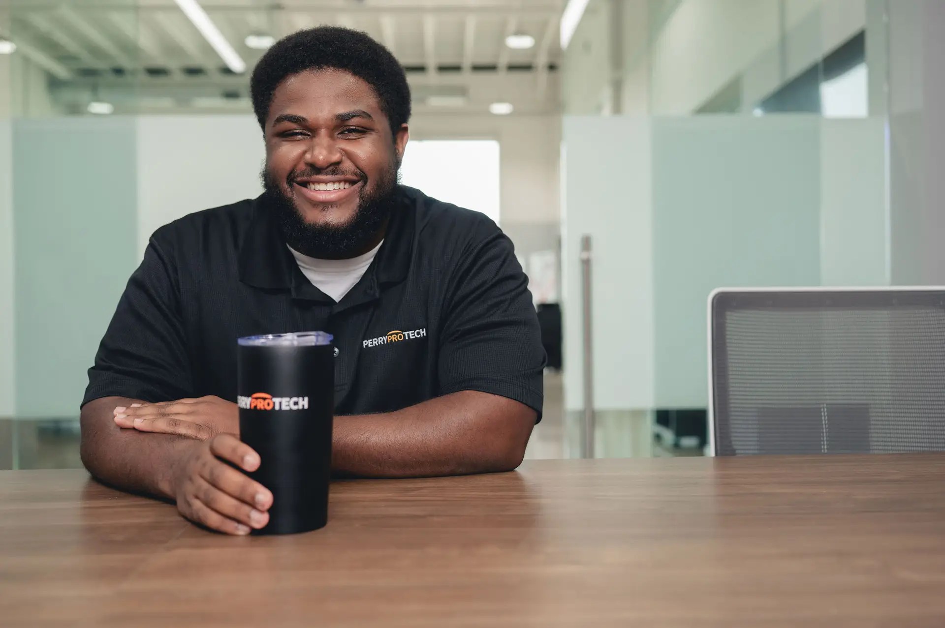 A smiling man with a beard, wearing a black shirt with a ProTECH logo, sits at a wooden table holding a black tumbler. The background is a modern office with glass walls and bright lighting.