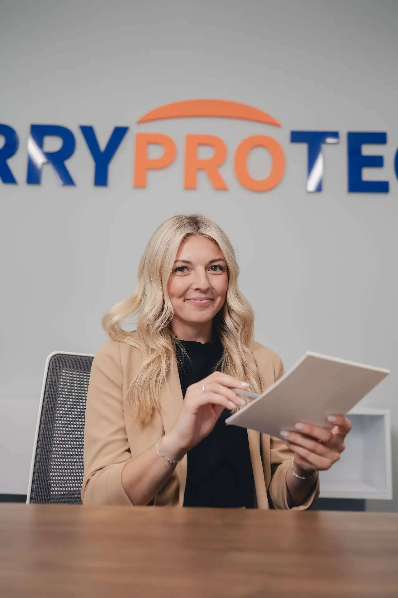 A woman with long blonde hair, wearing a tan blazer, sits at a desk holding a tablet. Behind her, a partially visible company logo hints at expertise in network security Ohio. She is smiling and facing the camera.