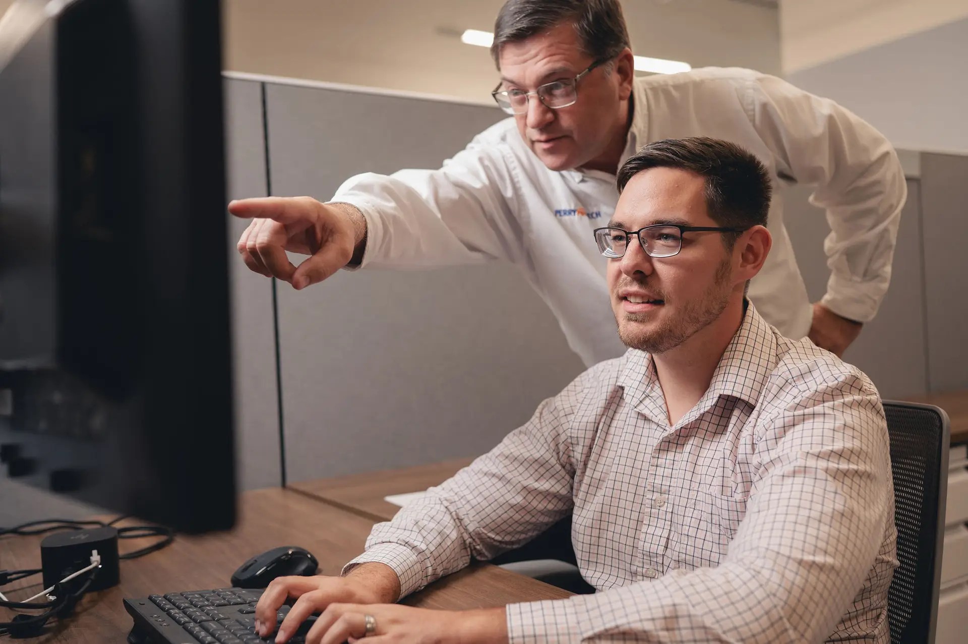 Two men in an office work at a computer; one sits and types while the other stands behind him, pointing at the monitor and giving guidance. Both wear glasses and business attire.