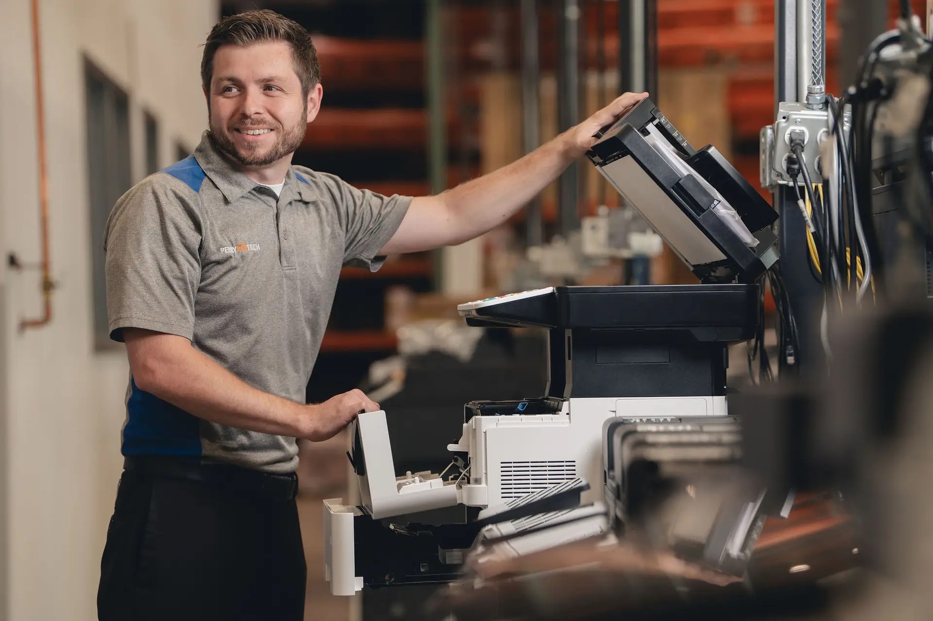 A smiling man in a gray polo shirt repairs or inspects a large office printer or copier in an indoor workspace, with shelves and equipment visible in the blurred background.