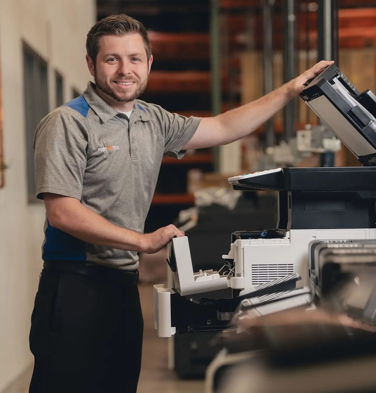 A smiling man in a gray polo shirt stands next to a large office printer, holding open a panel as if performing maintenance—demonstrating the reliability of Managed IT and Network Services in a brightly lit office environment.